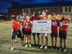 Winning soccer match team members standing on rec field, holding bracket sign, with CPASS building in the background. 