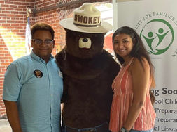 Malik Wynes, Smoky Bear, and Michaela Wynes pose for a photo in front of a Wynes Facility for Families and Children sign. 