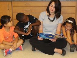 Female teacher wearing grey top and black pants reading a book to three young students, seated at her side. 