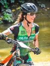Female mountain biker wearing helmet, gloves and sunglasses going through water, covered in mud.