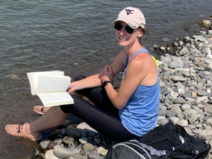 Catherine O'Malley sitting at the edge of the water reading a book with a flying WV hat on.