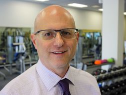 Peter Giacobbi is wearing a white dress shirt, dark tie and glasses, standing in front of work out equipment. 