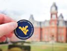 A hand holding a mountaineer coin in front of Woodburn Hall