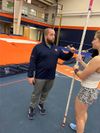 Coach wearing dark pullover talking with student athlete who is holding a pole vault.